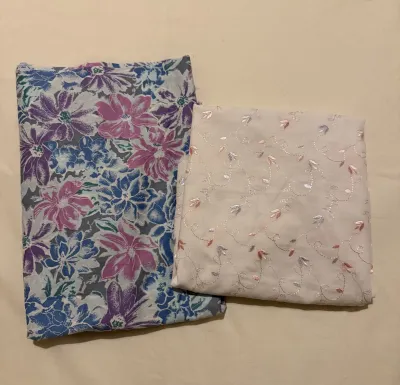 A white fabric background, on top are two pieces of folded fabric. One is grey, white, blue and purple florals and the other is white with lilac and pink embroidery flowers.