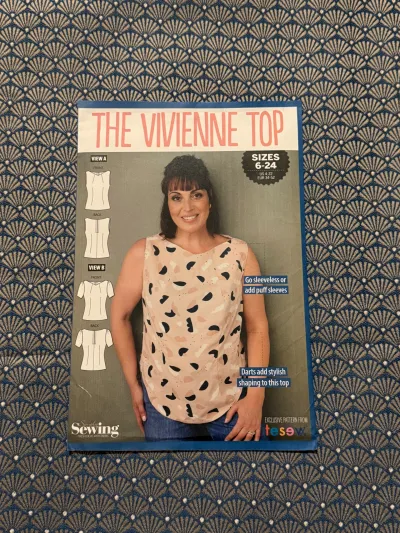 A blue fabric with small gold fans design, on top is a sewing pattern for the Vivienne top by Tesew in sizes 6-24. On the cover is a woman wearing a sleeveless top, next to her are drawings of a sleeveless top and a puff sleeve top.